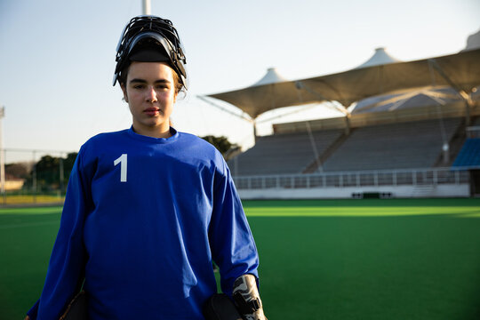 Field hockey goalkeeper standing on green turf pitch, wearing blue gear and padded gloves