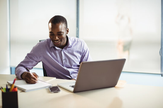 Pen writing notes in spiral notebook on office desk near window blinds with open laptop