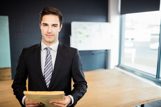 Businessman standing behind table in office, holding folder near whiteboard with charts and window