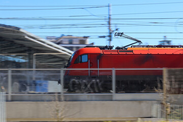 Modern red electric locomotive speeding through railway station with motion blur effect, overhead power lines visible against industrial urban setting