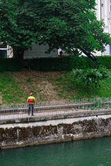 Man in a bright red and yellow jacket standing on a riverside path near lush green trees on a calm,...
