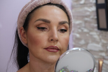 A young woman in home clothes carefully examines her fresh makeup in the mirror. Personal beauty routine, self-care, home comfort, natural beauty