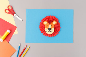 Colorful Lion paper craft project with red mane on blue sheet on grey background. Crafting tools include red scissors and yellow glue stick,