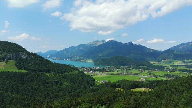 Drone ascending over Strobl town and lake Wolfgangsee in Austrian Alps, with forested mountains and bright sky with clouds.