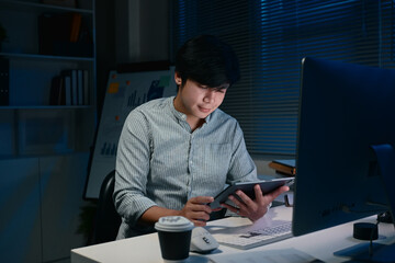 Young businessman working late at night in the office, using digital tablet under dim lighting
