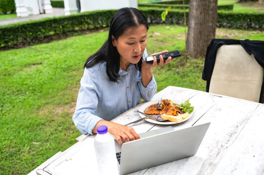 Busy Asian woman talking on phone and working on laptop during eating lunch in outdoor restaurant - Powered by Adobe