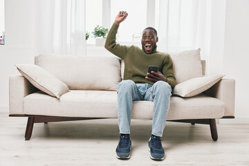 Happy young African American man celebrating success on his smartphone while sitting on a modern...