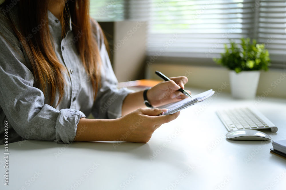 Poster close up of woman writing notes in a notebook while working at a desk with a computer keyboard.