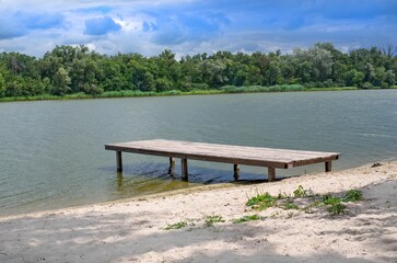 Peaceful riverside scene with a small wooden pier, sandy beach, calm water, and a forested...
