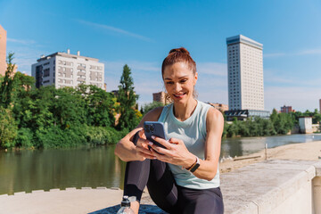 Smiling sportswoman reviewing fitness app near riverside, urban landscape gleaming with sunlight...