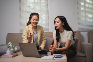 Two asian businesswomen are sitting on a sofa, discussing work and using a laptop in the living room, with one of them holding a pen and explaining something