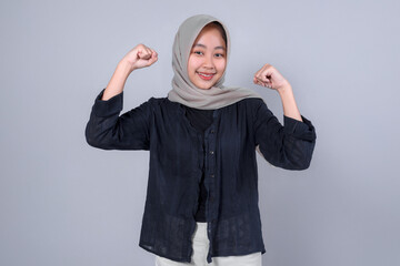 Confident young Indonesian Muslim woman wearing hijab and black shirt smiling while flexing her arms, expressing strength and empowerment on gray background.