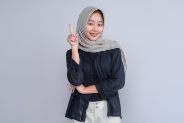 Cheerful young Indonesian Muslim woman in hijab smiling confidently while pointing upward with her index finger, symbolizing new ideas, awareness, solution, or inspiration.