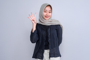 Portrait of a smiling young Indonesian woman in a hijab, showing four fingers with her right hand. She stands confidently against a plain grey background.