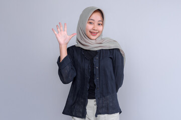 A cheerful young Indonesian woman in a light grey hijab and dark blouse smiles warmly while waving her hand, standing confidently against a plain light grey background.