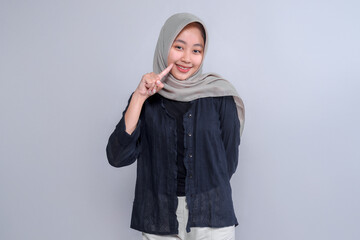 Portrait of a smiling young Indonesian woman wearing a grey hijab and black blouse, pointing to her cheek with one finger while standing confidently against a plain studio background.