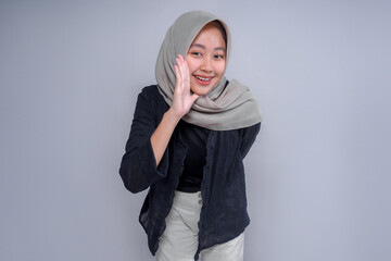 A cheerful Asian woman wearing a hijab and casual black attire, playfully cupping her hand to her...