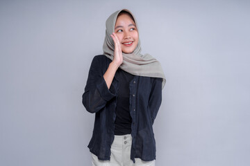 A cheerful Asian woman in a hijab, smiling and playfully cupping her hand to her mouth as if...