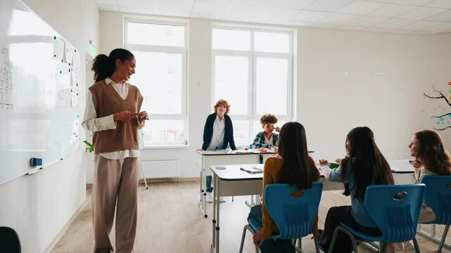 A female teacher is talking to a group of five schoolchildren sitting at desks while one of them stands