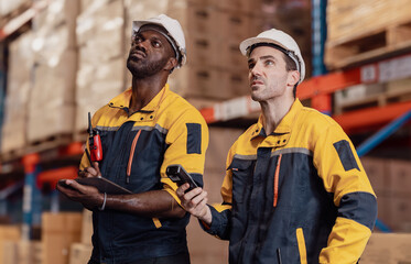 Fototapeta premium Warehouse manager, store inspector checking stock on storage shelf from inventory checklist. Two diverse ethnic people wear safety hard hat working together examining merchandise at distribution store