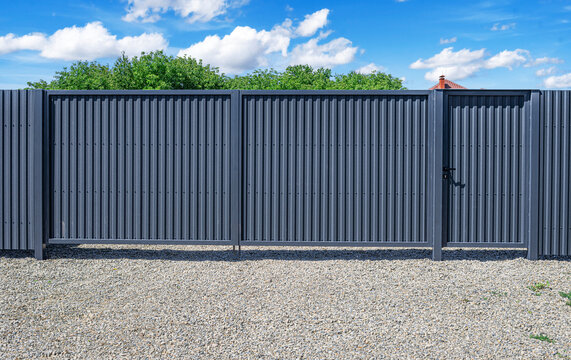 A modern gray corrugated metal fence with a built-in pedestrian gate stands on a gravel surface under a bright blue sky with scattered clouds.
