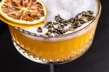 A macro shot of a foamy citrus cocktail topped with a dried lemon slice and scattered tea leaves,...