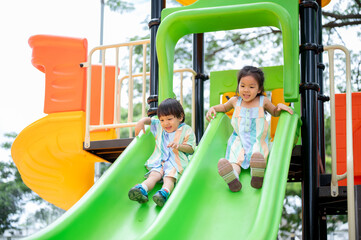 Asian girl and boy siblings having fun playing on colorful climbing slides in the park's playground.