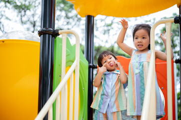 Asian girl and boy siblings making funny pose on colorful climbing slide in the park's playground.