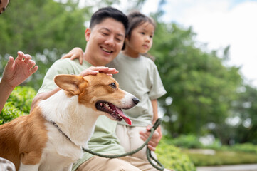 Smiling dad and daughter looking and patting corgi puppy while sitting on sidewalk bench in the park