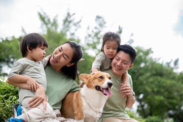 Mom looking at son with daughter hugging dad and patting corgi puppy while sitting on bench in park.