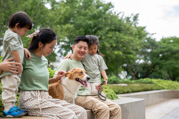 Smiling dad and daughter mother holding son patting corgi puppy head while sitting on bench in park.