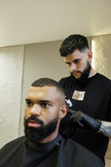 Fototapeta premium Tattooed barber trimming hair of african american client in barbershop