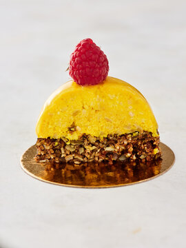 A vibrant yellow mousse dessert sits elegantly on a golden base, topped with a single, fresh raspberry. The dessert features a crumbly nut crust and showcases a tantalizing texture and vibrant color