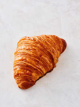 A warm, flaky croissant rests on a smooth, light-colored surface, showcasing its golden-brown layers. The pastry looks inviting and perfect for breakfast or a snack