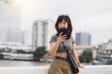 Happy young southeast asian woman teenager blogger using smartphone searching tourism guide. World travel explorer Asia concept for local cultural experience near Chao Phraya River Bangkok