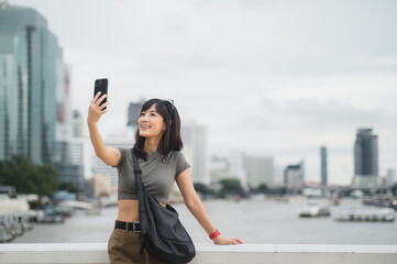 Beautiful young southeast asian woman blogger in 30s taking selfie with smartphone. World travel explorer local cultural experience summer tourism concept at Chao Phraya River Bangkok Asia