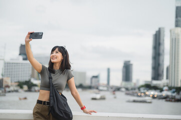 Beautiful young southeast asian woman blogger and world travel explorer in 30s taking happy selfie with smartphone at Chao Phraya River in Bangkok. summer tourism concept
