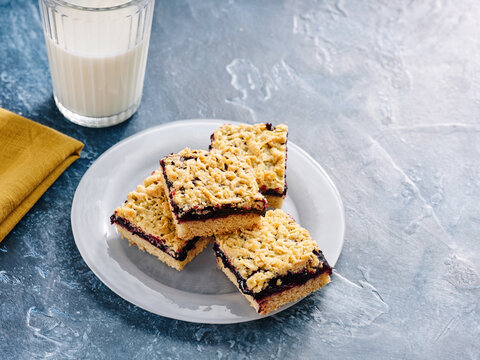 Four jam shortbread bars arranged on a white plate are paired with a glass of milk. The blue countertop adds a modern touch to this delightful snack setting