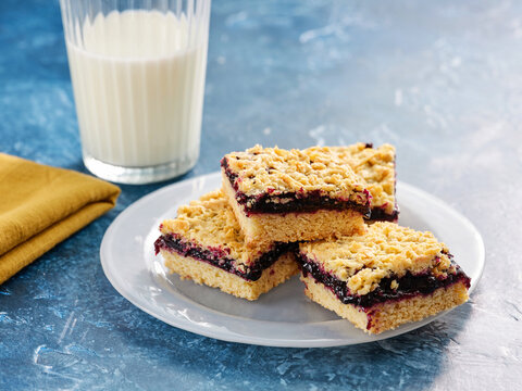 Soft and crumbly jam shortbread bars arranged on a white plate next to a glass of milk. The treat features a fruity layer and a golden crumb topping, perfect for any snack time