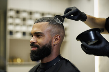 Barber applying hair dye on head of smiling african american client in barbershop