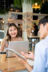Asian woman looking at a man holding tablet talking as sits with laptop at table in cafe or canteen