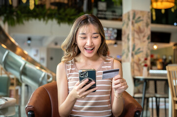 Asian woman holding phone looking happily at credit card as sitting on brown sofa in cafe or canteen