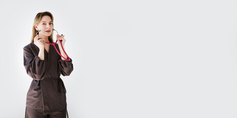 Wide banner with young female doctor in dark uniform holding a pink stethoscope on light background with copy space for text or design.