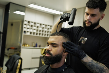 Tattooed barber drying hair of african american man after haircut in barbershop