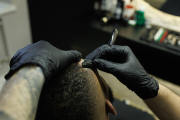 Tattooed barber styling hair of client with razor in barbershop 