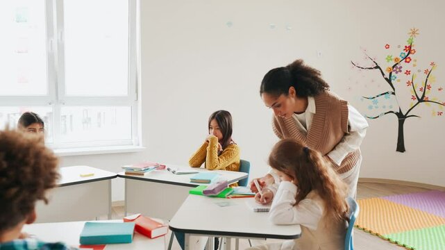 Female teacher holding cards and writing in notebook to schoolgirl sitting at desk near group of three classmates
