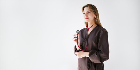 Wide banner with young female doctor in dark uniform using a pink stethoscope, looking aside, on light background with copy space for text or design.