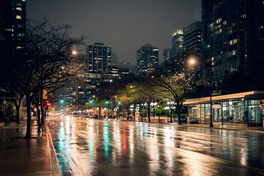 Rainy city street at night with glowing reflections on wet pavement, modern buildings and warm streetlights, moody blue and yellow palette, with space for text. - Powered by Adobe