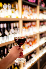 Close-up of a hand holding a glass of red wine in a liquor store with blurred shelves of wine bottles in the background. Elegant lifestyle and wine appreciation concept.
