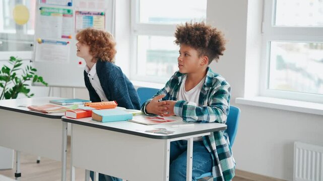 Two schoolchildren are sitting at desks and talking while one of them gets up and sits on a chair